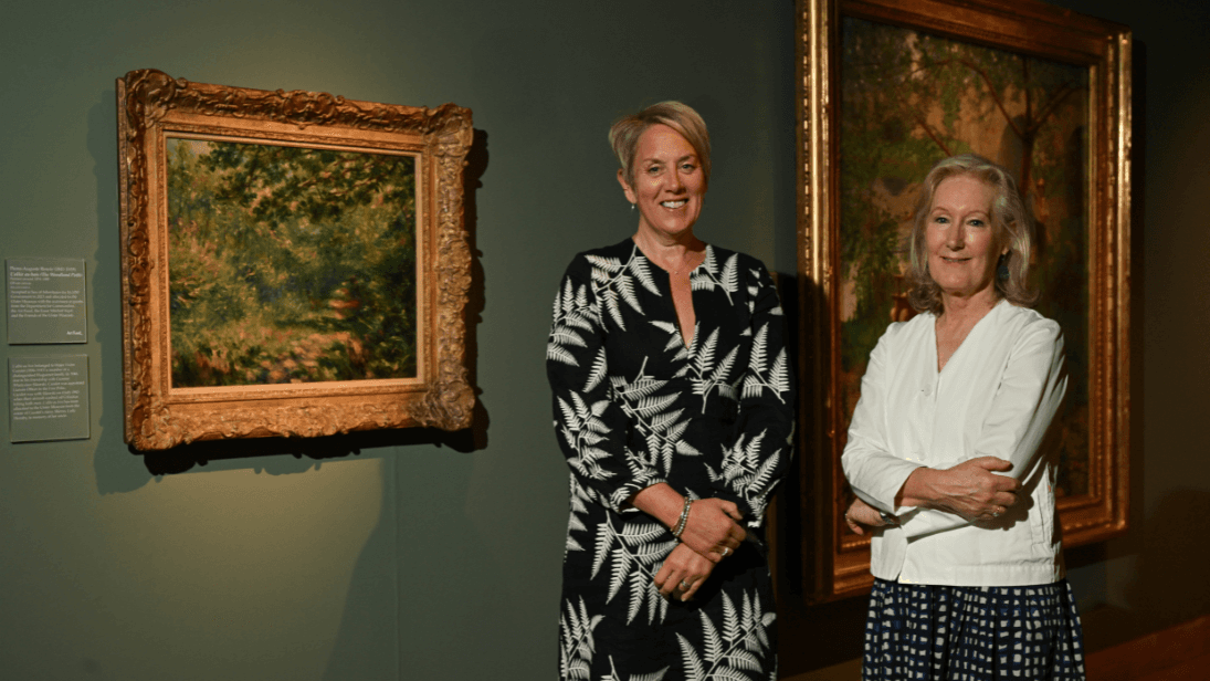 Renoir's L’allée au bois hung on a green wall with dimmed light shining down on the painting, with the Chief Executive of National Museums NI, Kathryn Thomson and Senior Curator of Art, Anne Stewart standing in front of it.