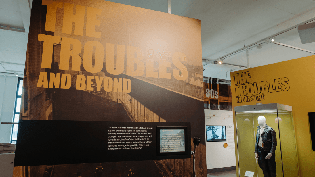 Inside the Troubles and Beyond exhibition at Ulster Museum. Yellow and black graphics on the wall say 'The Troubles and Beyond' and there is a glass display case in the background with a jacket artefact.