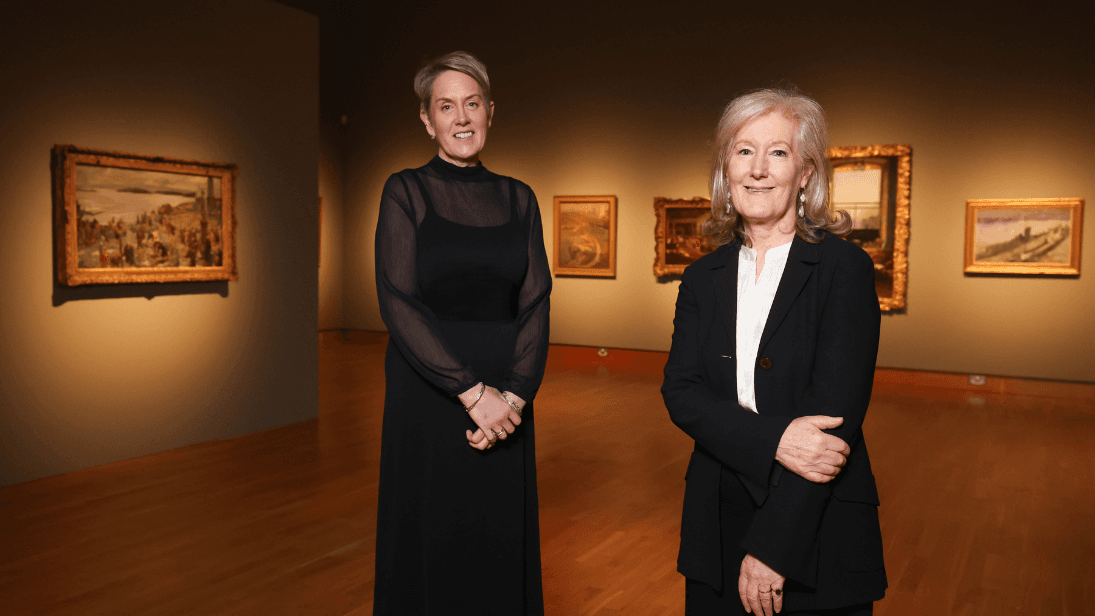 Two women dressed in black outfits standing in front of paintings by Sir John Lavery.