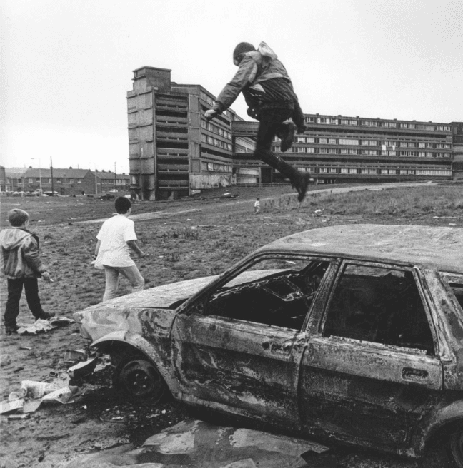 Original photographic print showing a boy leaping off a burnt-out car at Divis Flats