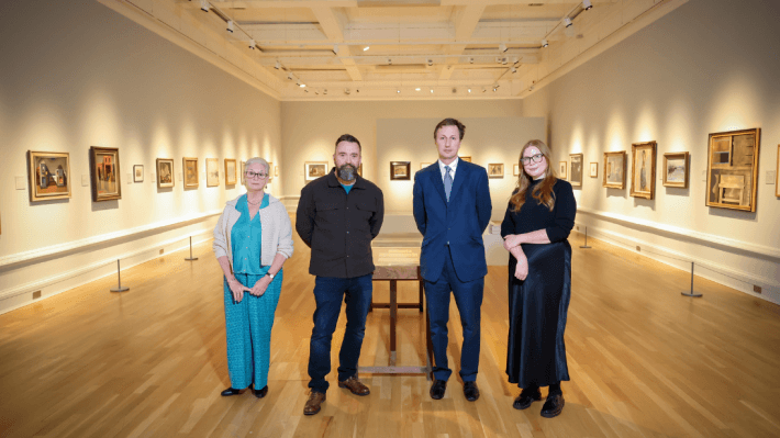 Four people standing in a lit up gallery, with paintings on every visible wall, at the opening of an exhibition at Ulster Museum.