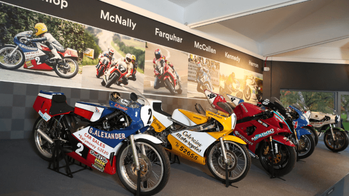 Six motorcycles displayed on a grey slanted platform with pictures of the racers that rode them on wallpaper on the wall behind each one. From left to right the wallpaper says Dunlop, McNally, Farquhar, McCallen, Kennedy and Herron.